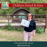 A smiley Give a Book member, Ali, stands in front of a wooden 'hobbit hut', holding a copy of Michael Rosen's, 'We're Going on a Bear Hunt'.