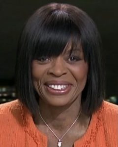 Ronke Phillips, in a black top and orange blazer, sat at a news desk in front of a backdrop of London, lit up at night