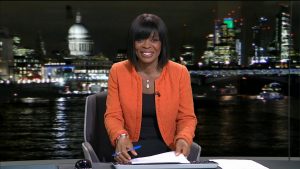 Ronke Phillips, in a black top and orange blazer, sat at a news desk in front of a backdrop of London, lit up at night